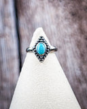 The Bahman Adjustable Genuine Turquoise Ring on a display stand with a wood backdrop.