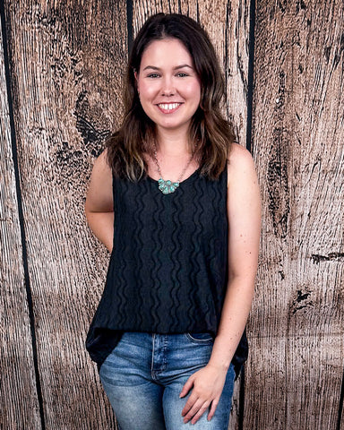 The Caldera Tank in Black being styled with jeans and modeled by a women in front of wood backdrop.