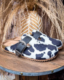 Andi Slip on Mule in Black Cow Print by Very G displayed on wooden table in front of wheat backdrop.