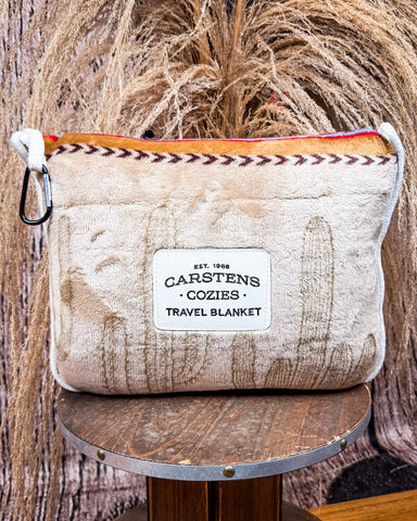 Front view of the bagged Cactus Stripe Travel Blanket on a wooden table with wheat behind it.