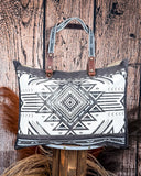 The Black Sands Weekender Bag displayed on a wooden table with wheat in the back, wood backdrop.