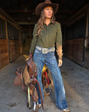 Woman in cowboy hat and jeans standing in a stable with a saddle.