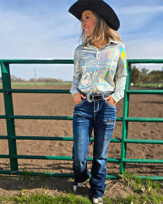A woman wearing a shiny silver long-sleeve button-up shirt, blue jeans, a black cowboy hat, and a black belt stands outdoors in front of a green metal fence and dirt field.