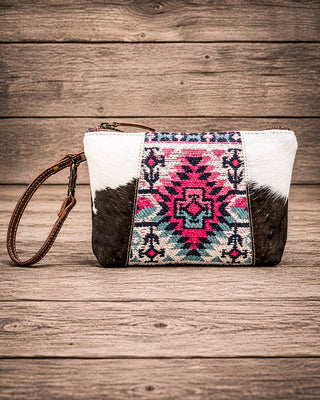 A western-style clutch bag with a cowhide pattern and a central geometric Southwestern-style embroidered panel, featuring a brown leather wristlet strap, set against a rustic wooden plank background.