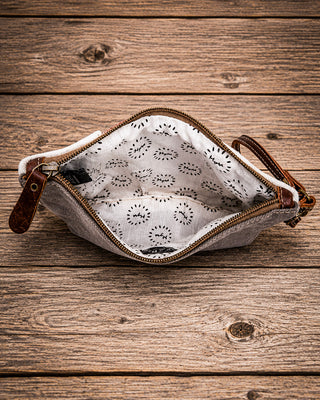 An open canvas zippered pouch with a brown leather zipper pull and handle, showing a white patterned interior, set on a rustic wooden plank background.