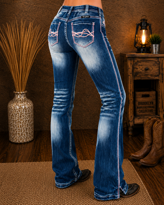 Rear view of a person wearing blue western flare jeans with white contrast stitching and decorative stitching on the back pockets, standing in a rustic room with cowboy boots, a wooden cabinet, and a lamp in the background.