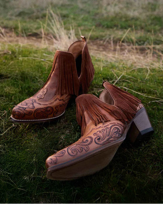 A pair of brown leather cowboy boots with fringe and tooling details, resting on green grass in a field with cows in the background.