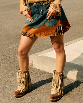 Person wearing a distressed top, denim skirt, and fringed boots in an outdoor setting.