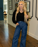 Woman wearing a black top and blue jeans indoors with framed pictures on the wall.