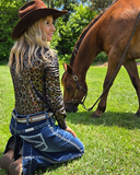 Woman in cowboy hat and leopard print shirt kneeling next to a horse on grass