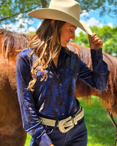Woman in a blue shirt and cowboy hat standing next to a horse outdoors.