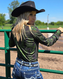 Woman in a cowboy hat and green long-sleeve shirt standing next to a green fence with trees in the background.