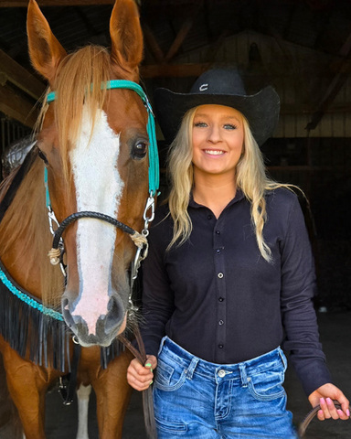 Woman in a black shirt and blue jeans standing next to a brown horse in a stable.