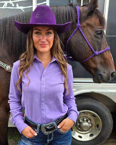 Woman in a purple shirt and cowboy hat standing next to a horse with a purple halter.