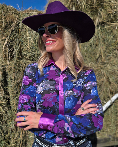 Woman wearing a purple floral shirt and wide-brimmed hat standing in front of hay bales.