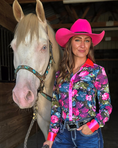 Woman in a colorful floral jacket and pink cowboy hat standing next to a horse in a barn.