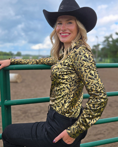 Woman wearing a gold and black patterned shirt and black cowboy hat, standing in an outdoor setting.