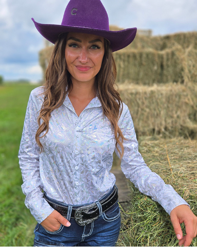 Woman wearing a purple cowboy hat and white long-sleeve shirt in a field with hay bales.