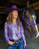Woman in a purple iridescent shirt and cowboy hat standing next to a brown horse with a purple halter in a barn.