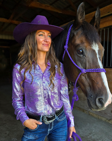 Woman in a purple iridescent shirt and cowboy hat standing next to a brown horse with a purple halter in a barn.