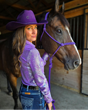 Woman in a purple shirt and cowboy hat standing next to a horse in a stable.