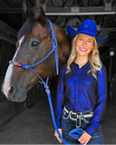 Woman in blue glitter shirt and cowboy hat standing next to a horse in a stable.