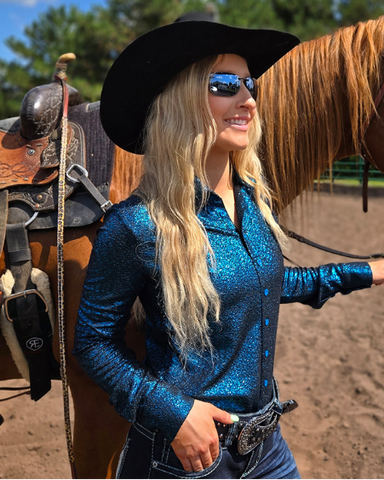 Woman in a teal glittery shirt and black cowboy hat standing next to a horse in an outdoor setting.