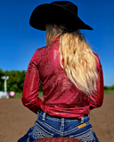 Person wearing a red foil shirt and black cowboy hat, standing on a dirt arena with a blue sky.
