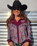 Woman wearing a patterned shirt and cowboy hat with sunglasses, standing against a horse trailer.