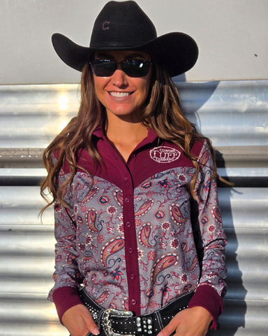 Woman wearing a patterned shirt and cowboy hat with sunglasses, standing against a horse trailer.
