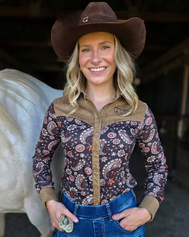 Woman in cowboy hat and patterned shirt standing next to a horse in a stable.
