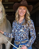 Woman in a cowboy hat and patterned shirt standing next to a horse in a stable.