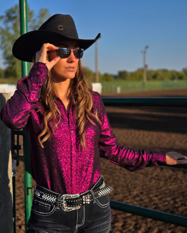Woman in a magenta glittery shirt and black cowboy hat adjusting sunglasses outdoors.