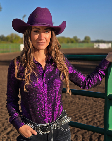 Woman in a purple glitter shirt and cowboy hat standing in a dirt arena.
