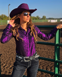 Woman in a purple glittery shirt and cowboy hat standing in a dirt field.