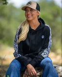 Woman wearing a black hoodie with patterned sleeves and a beige cap, sitting outdoors.