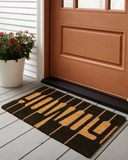 Howdy Coir Doormat in front of a brown door on a wooden floor.