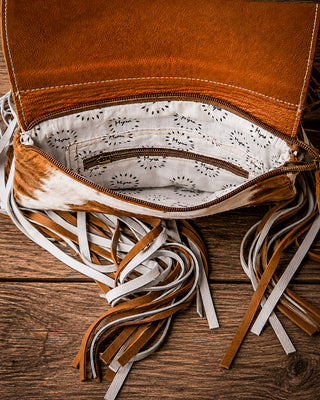 A close-up of a brown leather fringe bag shown open, revealing a patterned cream fabric lining, placed on a rustic wooden surface.