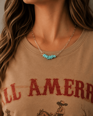 A close-up of a woman wearing a silver chain necklace with a row of turquoise-colored beads at the center, paired with a beige graphic t-shirt.
