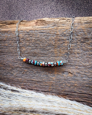 A silver chain necklace with a curved bar of colorful marbled beads, displayed on a rustic wooden log with dried grass in the foreground.
