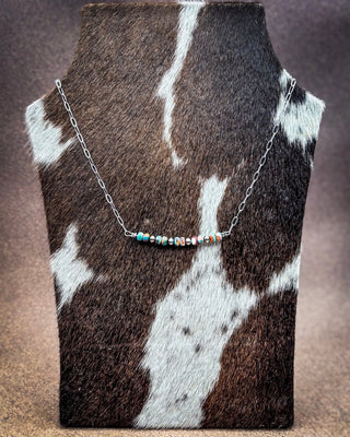A silver chain necklace with a central cluster of small colorful beads, displayed on a cowhide jewelry bust against a brown textured background.
