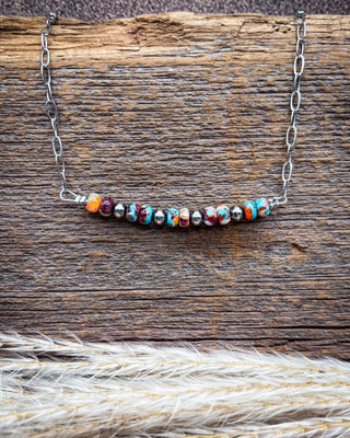 A silver chain necklace with a curved bar of colorful marbled beads, displayed on a rustic wooden surface with dried grass in the foreground.