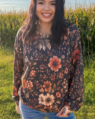 A smiling woman with long dark hair stands outdoors in a grassy field with a cornstalks background, wearing a dark brown long-sleeve top with a red, orange, and beige floral pattern, paired with blue jeans.