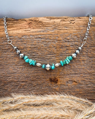 A silver-toned beaded necklace with turquoise and metallic beads, displayed on a rustic wooden surface with dried grass in the foreground.