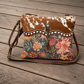 A western-style crossbody bag with a brown and white cowhide-patterned top flap, a decorative scalloped trim, and a colorful floral print body, displayed on a rustic wooden plank surface.