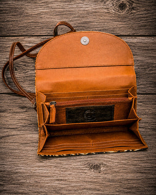 An open tan leather wallet or clutch with multiple interior card slots, a zip pocket, and a shoulder strap, photographed on a rustic wooden surface.