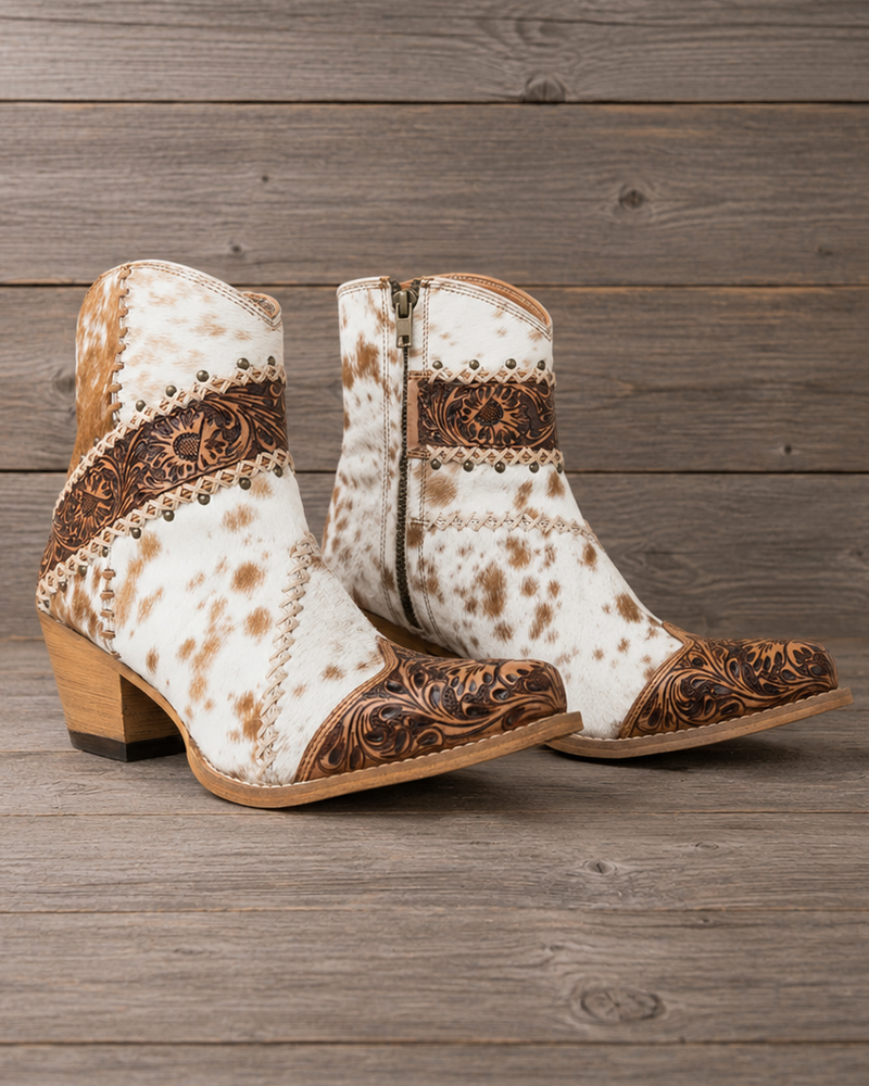A pair of women's western ankle boots with a white and brown cowhide pattern, brown tooled leather detailing, and stacked wooden block heels, displayed on a rustic gray wood plank background.