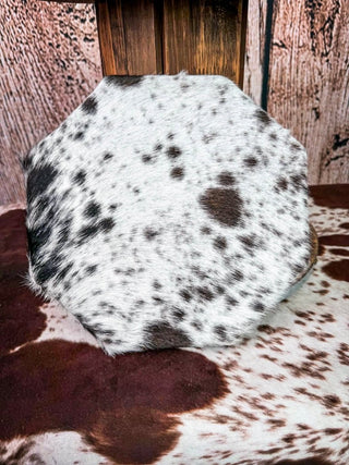 An octagonal decorative tray or bowl with a dark brown leather rim and a white cowhide center featuring dark brown spots, displayed on a cowhide rug in front of a rustic wood and leather backdrop.
