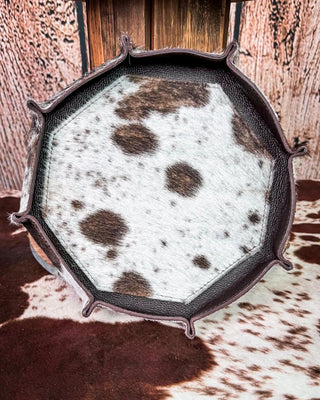 An octagonal decorative tray or bowl with a dark brown leather rim and a white cowhide center featuring dark brown spots, displayed on a cowhide rug in front of a rustic wood and leather backdrop.