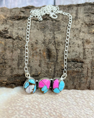 A silver chain necklace with three oval turquoise and pink stone pendants, displayed on a textured wood surface with a light fabric background.